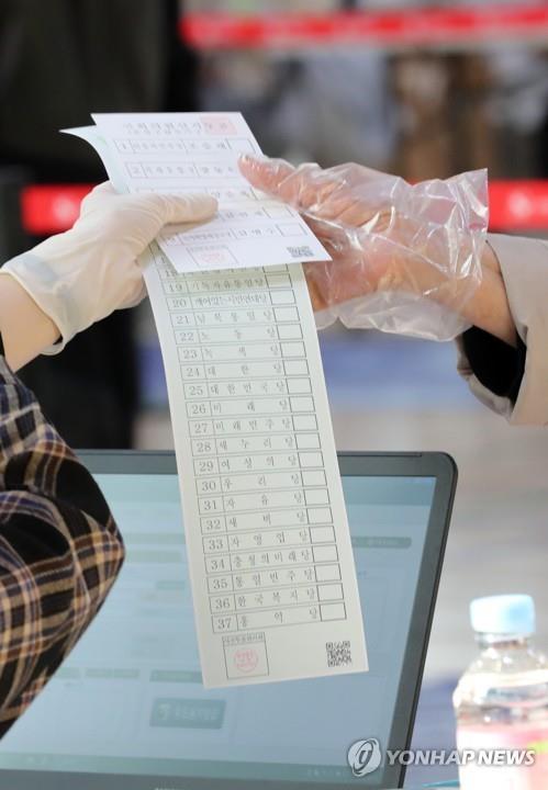 2020년 4월 10일 대전 유성구 온천2동 사전투표소가 마련된 어은중학교 다목적강당에서 유권자가 장갑을 끼고 투표지를 받고 있다. 한국은 후보자가 많으면 투표 용지가 길어진다. 반면 도쿄 지사 선거는 투표용지에 후보자의 이름을 인쇄하지 않고 유권자가 필기구로 직접 적는 방식이라서 후보가 많았지만, 투표용지 크기는 작았다. [연합뉴스 자료사진]