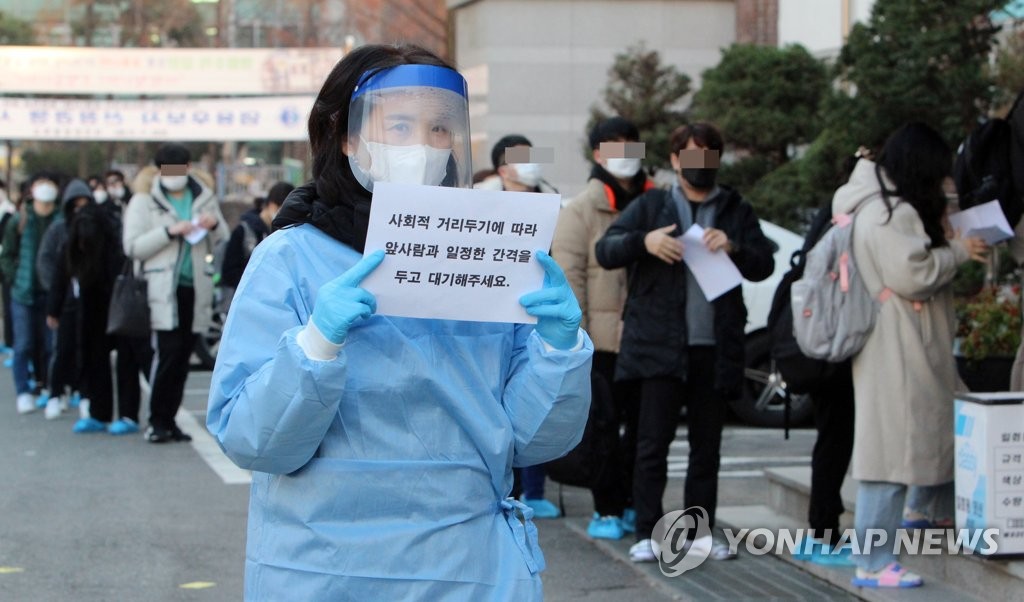 An official holds up a sign on coronavirus prevention guidelines as applicants wait in line to enter a test site for a state-run teacher employment examination in the central city of Daejeon on Nov. 21, 2020. (Yonhap)